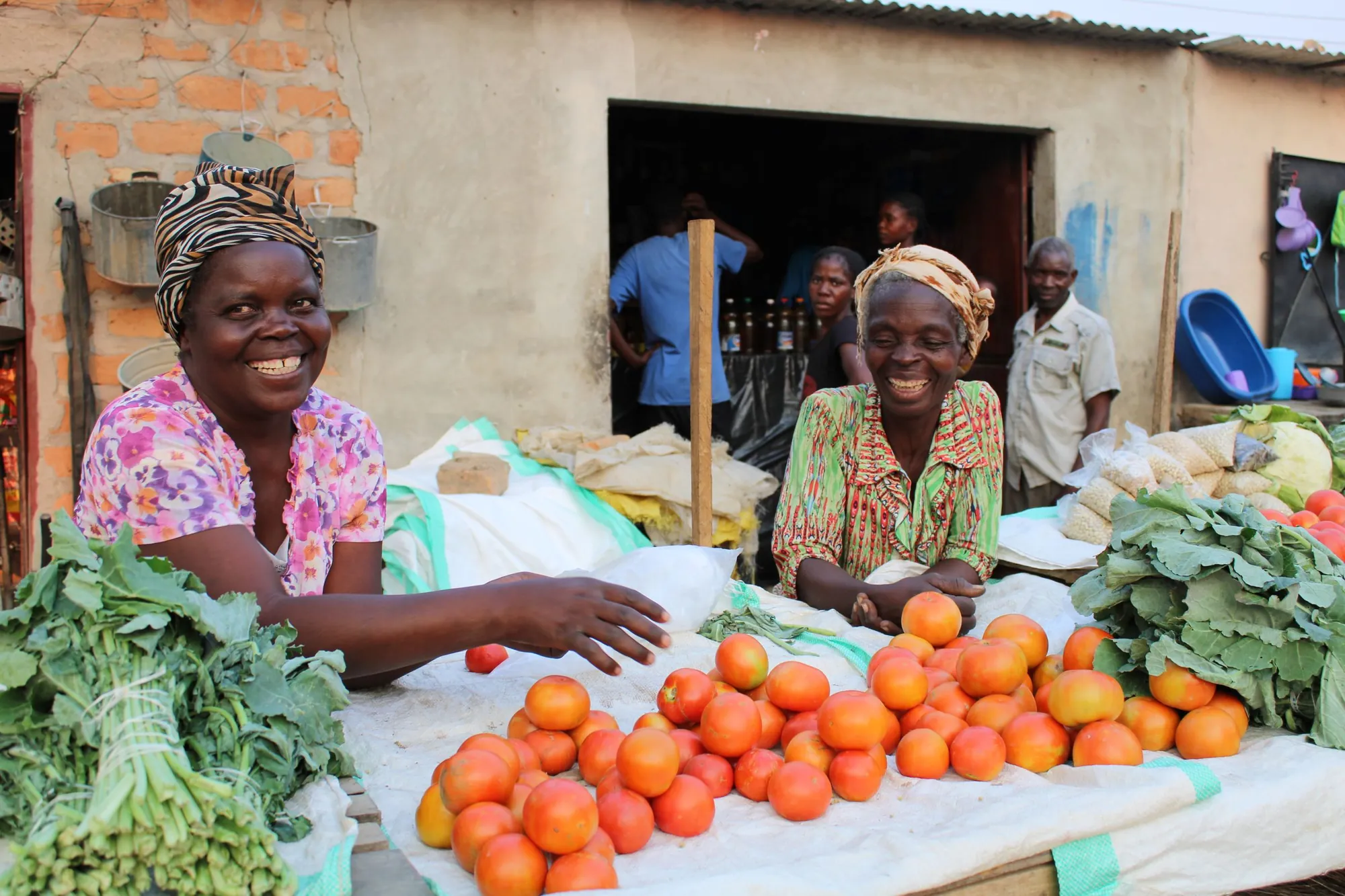 Two women laughing at their produce stall, with tomatoes and greens laid out in front of them